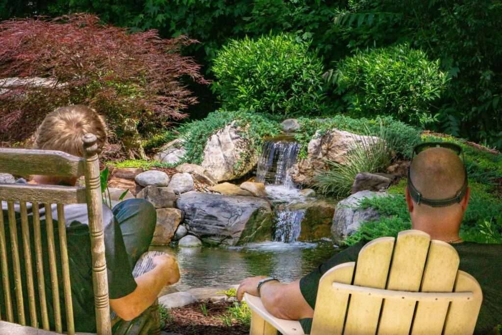 Greg Wittstock, also known as “The Pond Guy,” sitting in a chair with Bradley Blocksom enjoying a custom-built backyard waterfall surrounded by natural landscaping in Weddington, North Carolina