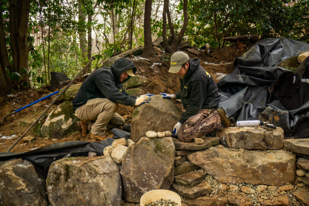 Pond builders positioning boulder during natural pond installation in Matthews, NC