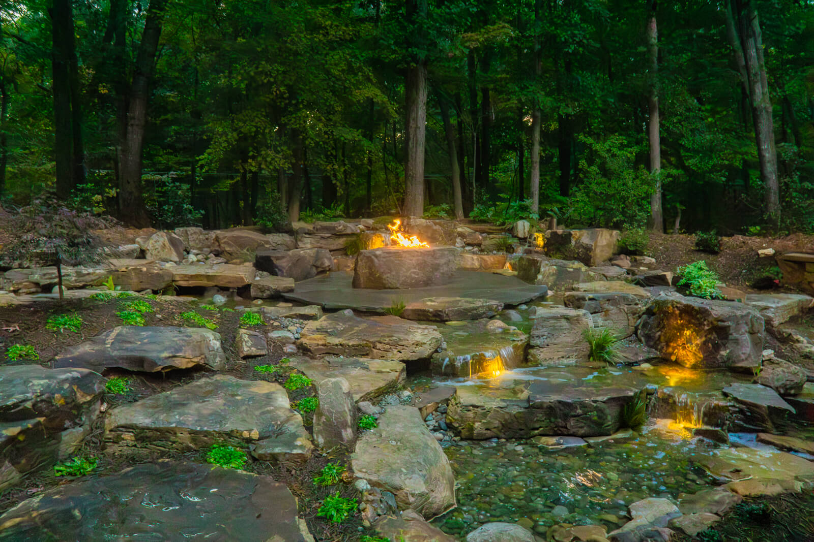Custom pondless waterfall with stepping stones, integrated fire pit, and waterfall lighting in Weddington NC