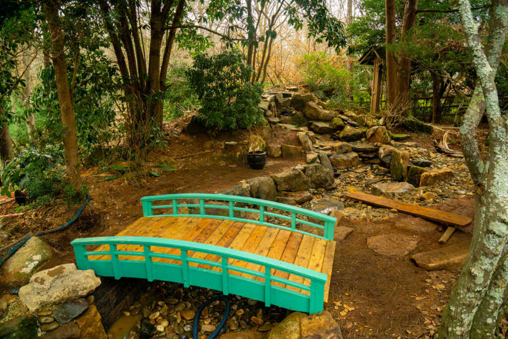 Bright blue garden bridge installed over stream style pond with garden path Matthews NC