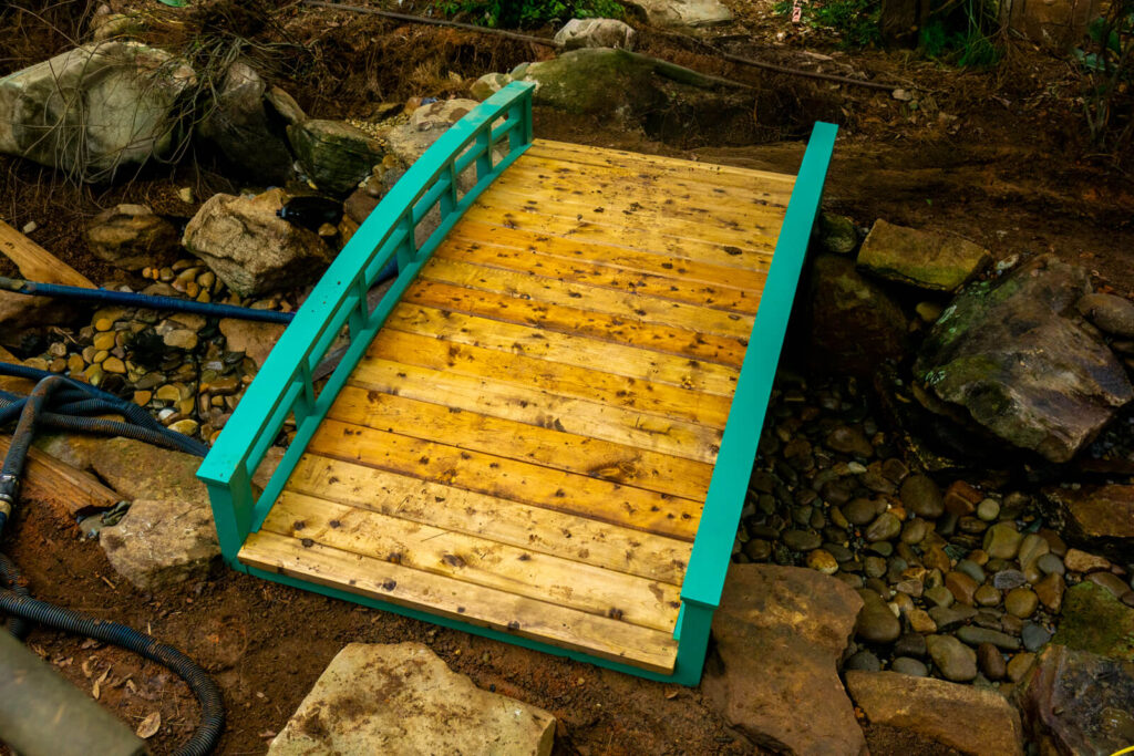 Close up of bright blue wooden garden bridge spanning stream pond during installation Matthews NC