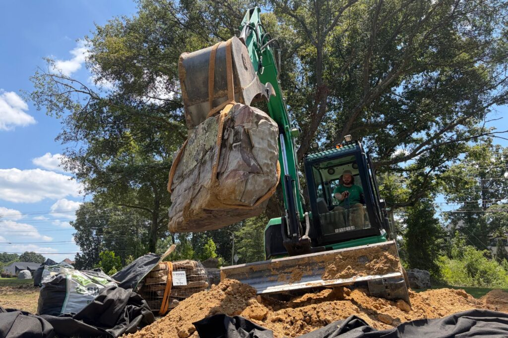 Excavator carefully transports a large boulder suspended by lifting straps above the pond liner.