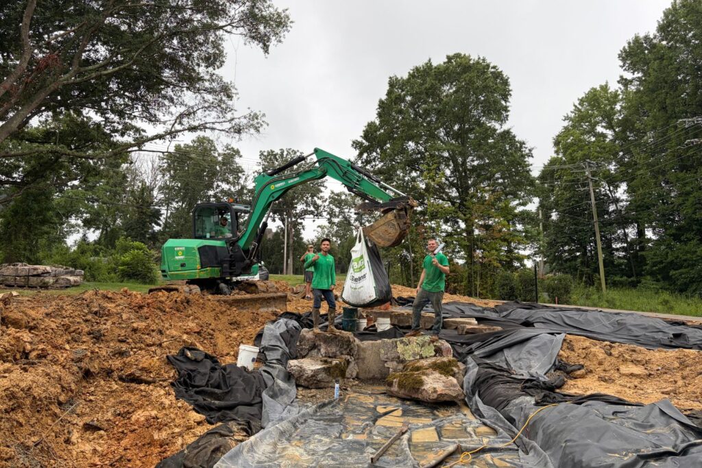 Landscape construction team installs gravel and river rock in a custom ecosystem pond using an excavator.