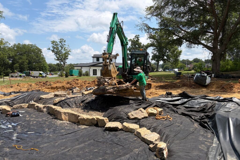 Construction crew uses an excavator to place large natural boulders within a pond excavation.