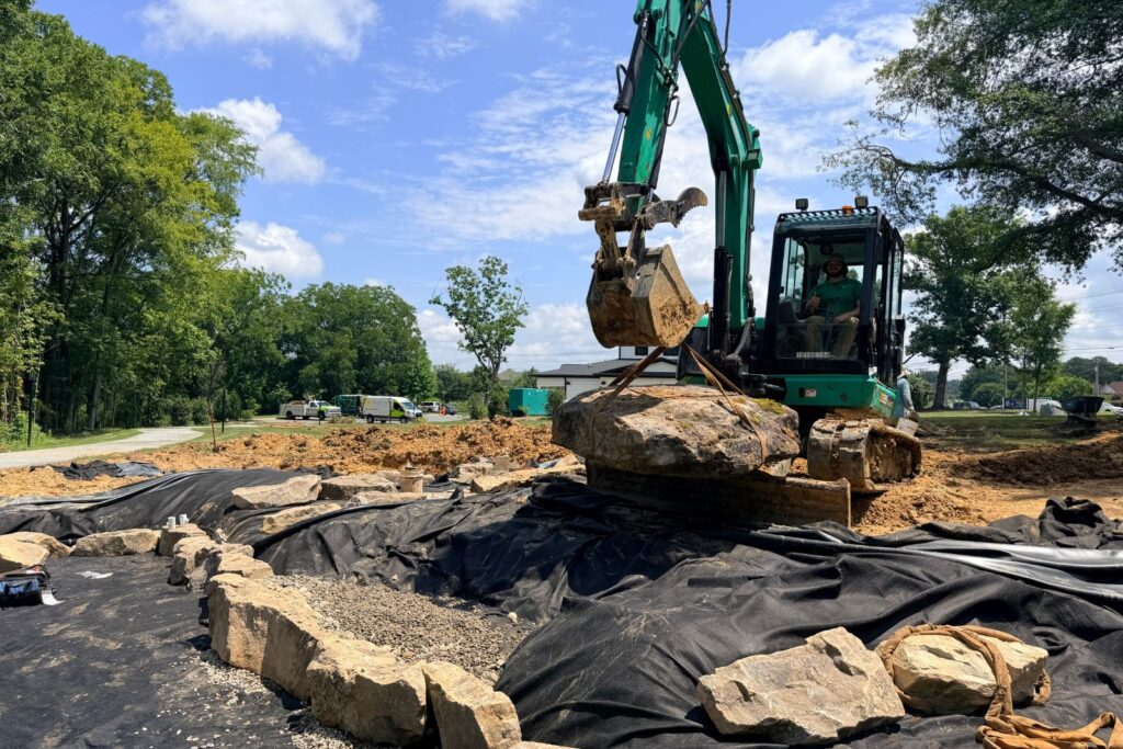 Excavator places a large natural stone boulder into a lined pond excavation during the construction of a custom water feature in Houston Ross Park.