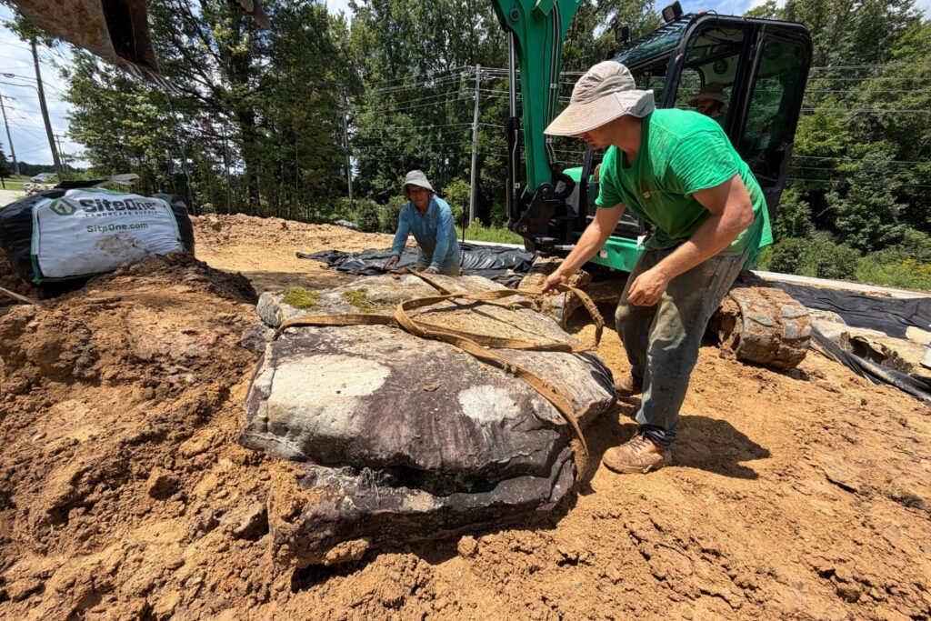 Crew member lifting straps around a large natural boulder beside an excavator.