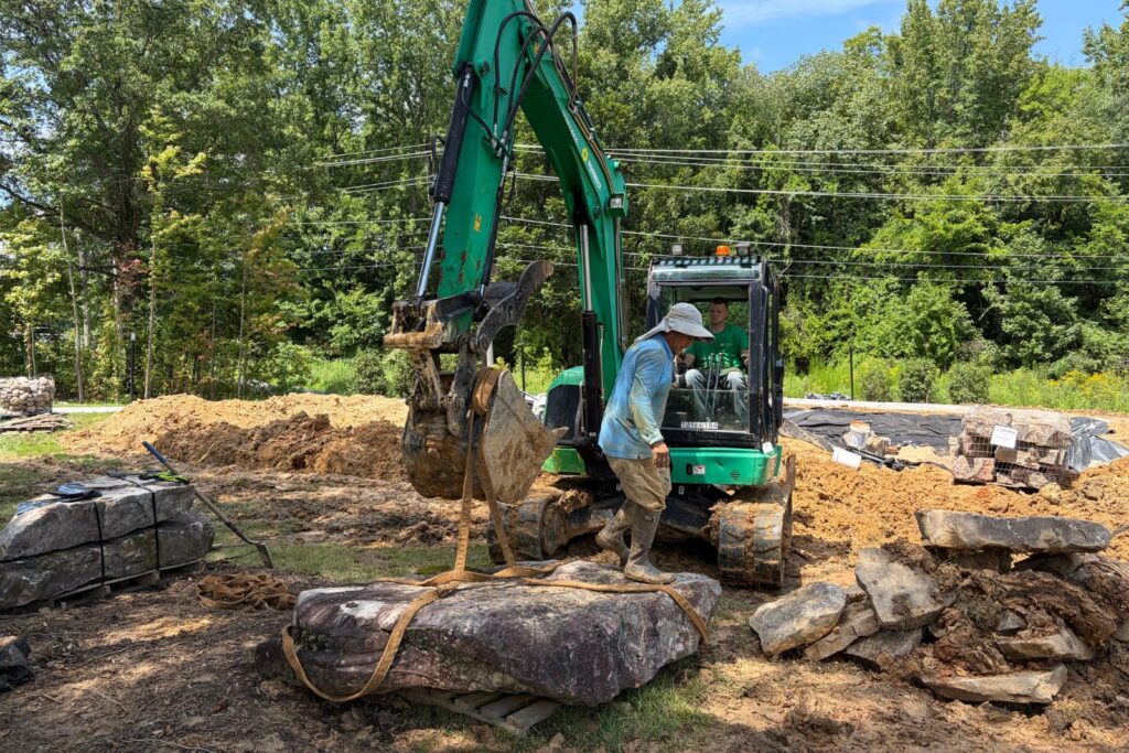 Excavator and construction crew member maneuver a large moss-covered boulder into position.