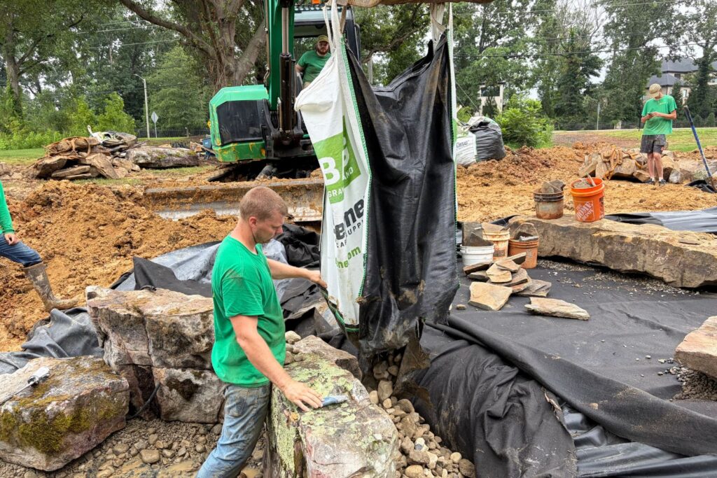 Crew member installs gravel and river rock around large decorative boulders inside a lined pond basin.
