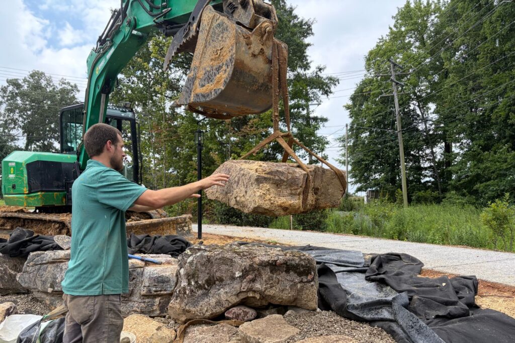Crew member guides a suspended natural stone boulder into position with lifting straps and an excavator.