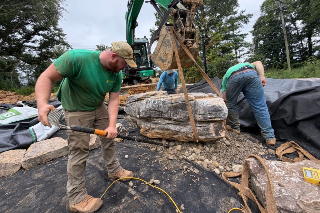 Crew levels drainage gravel and carefully sets a large natural stone slab inside a lined pond basin.