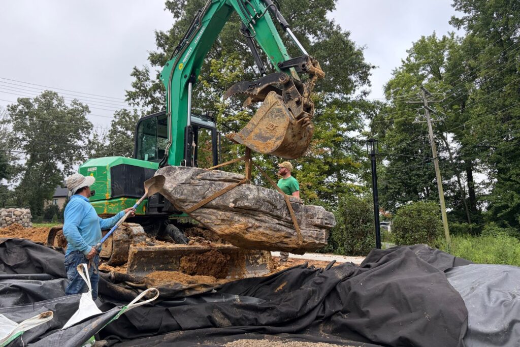 Excavator lifts and maneuvers a natural boulder into place during custom pond and waterfall construction.