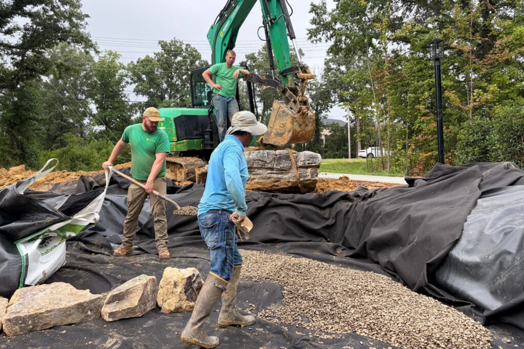 Construction crew spreads drainage gravel and positions large natural stone boulders inside a lined ecosystem pond excavation.