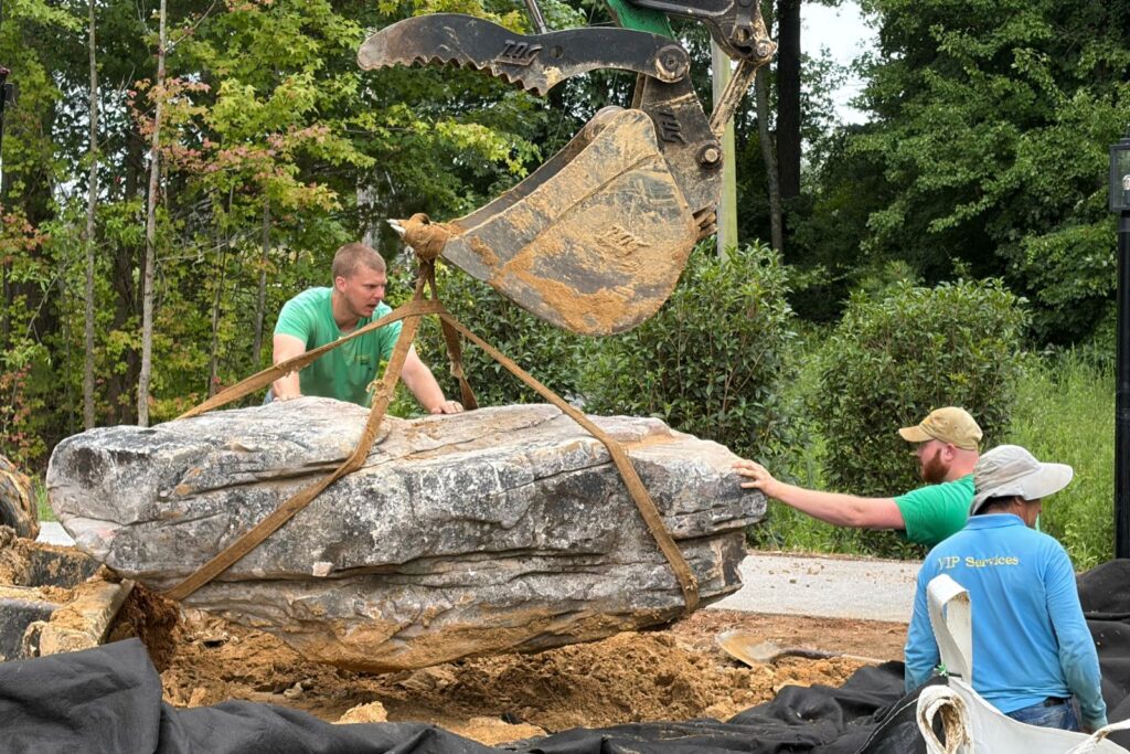 Crew guides a suspended natural stone into position using an excavator.