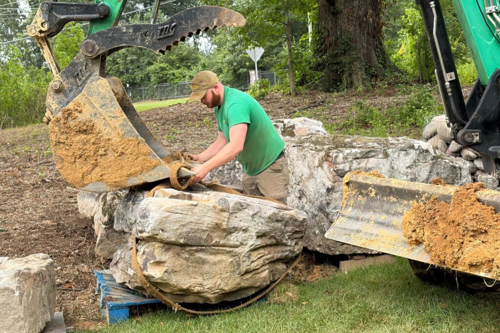 Crew member secures lifting straps around a large boulder beside an excavator, preparing natural stone for placement in a custom rainwater harvesting system installation.