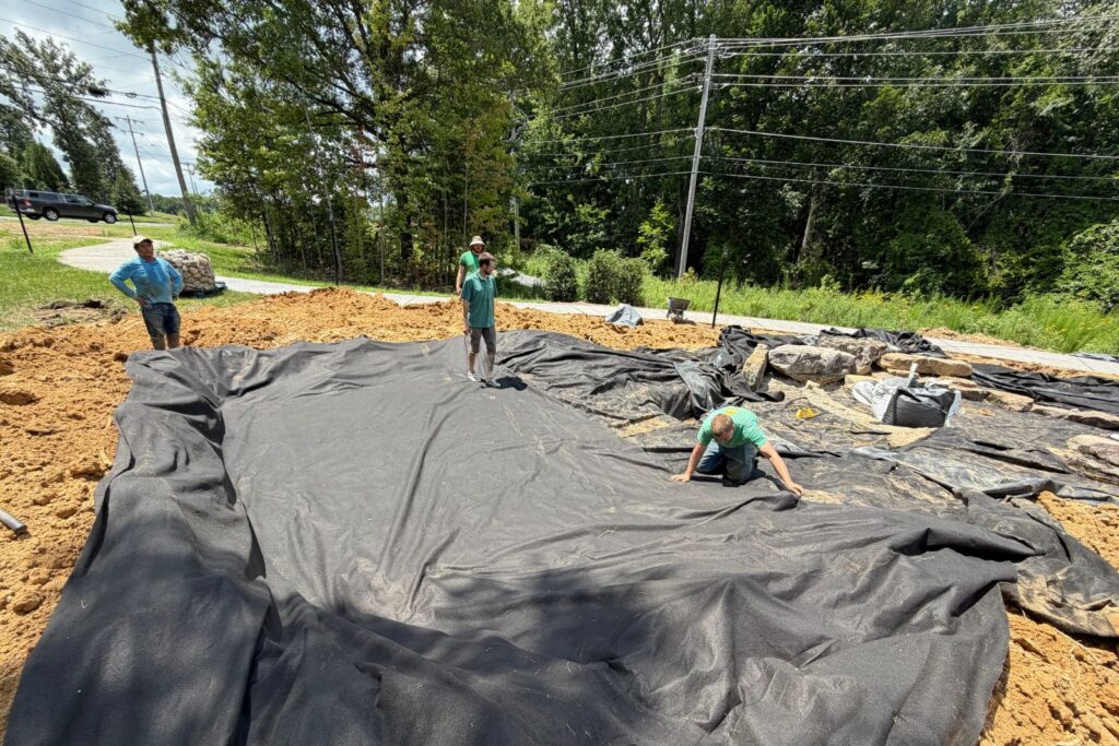 Construction crew installs protective underlayment fabric across a large pond basin, preparing the foundation for new water garden feature in Marvin, NC.
