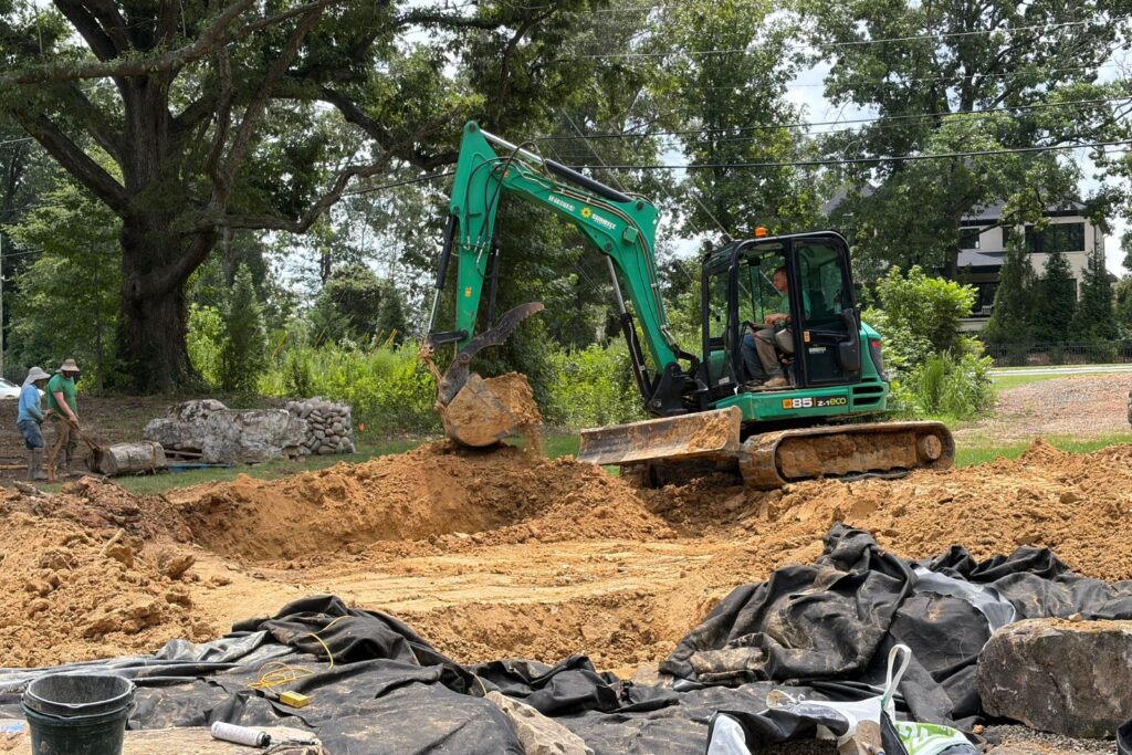 Excavator performs grading and earthmoving work around a custom pond excavation, shaping the basin and waterfall areas.